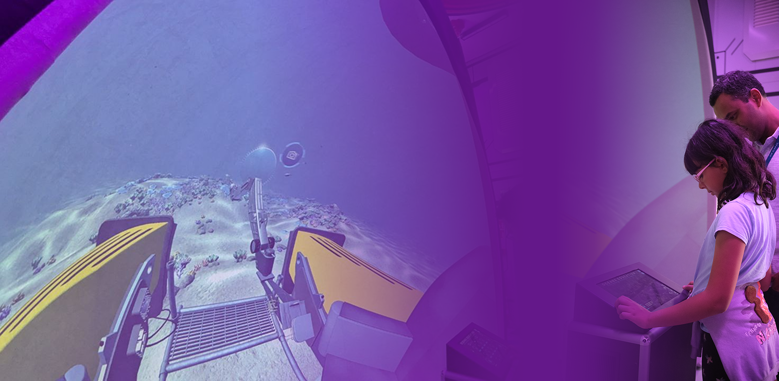 A child and parent looking into the Dive & Discover dome simulation, showing the submersible's cockpit view of a coral reef, shown in a duotone treatment.